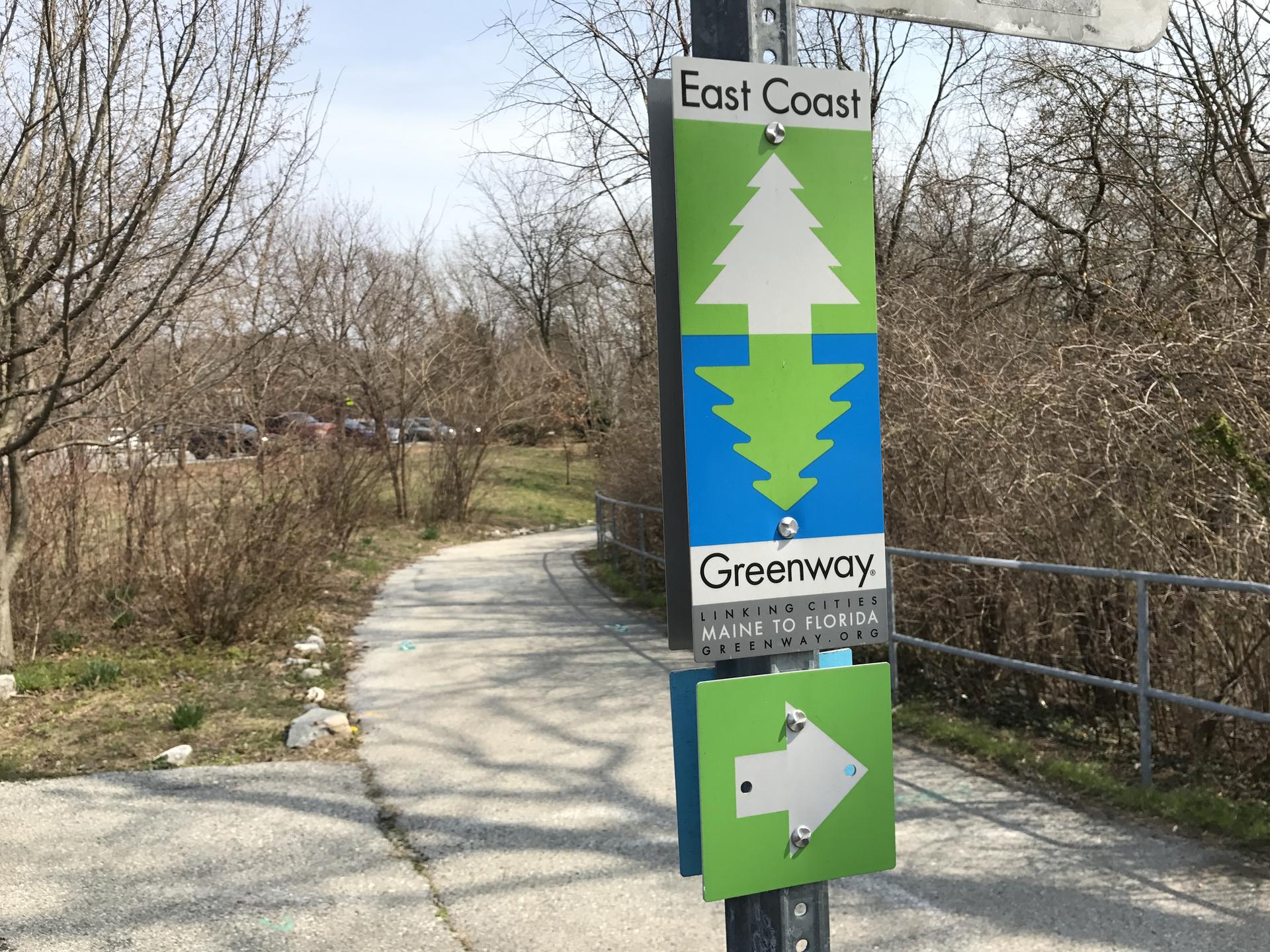 A green trail sign stands beside a paved path through bare trees.