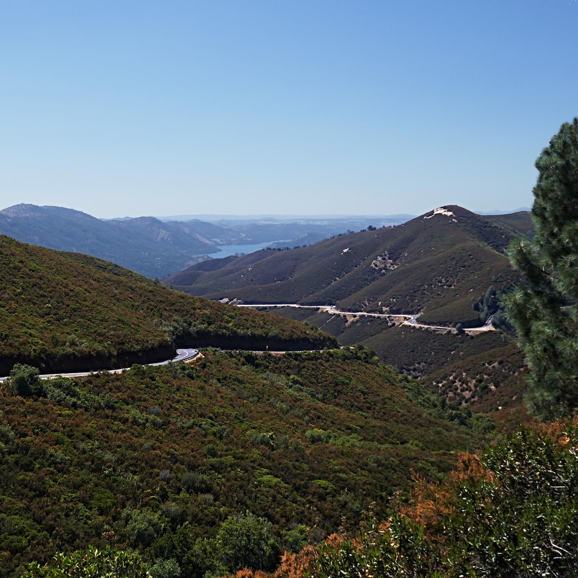 Winding road through green, sunny mountains with a distant lake under clear sky.