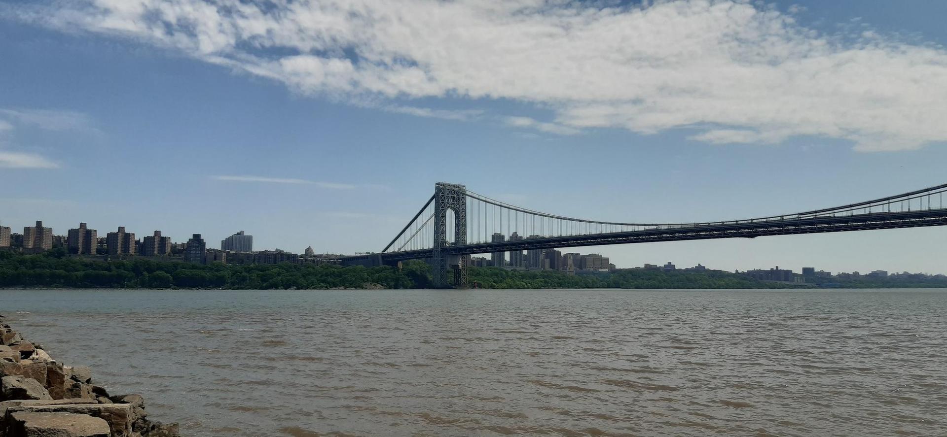 Suspension bridge spans a wide river with a distant city under blue sky.