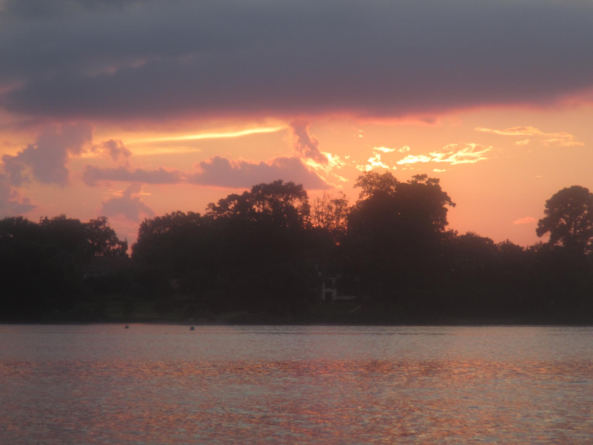 Vibrant sunset with golden and purple clouds over a calm lake and silhouetted trees.
