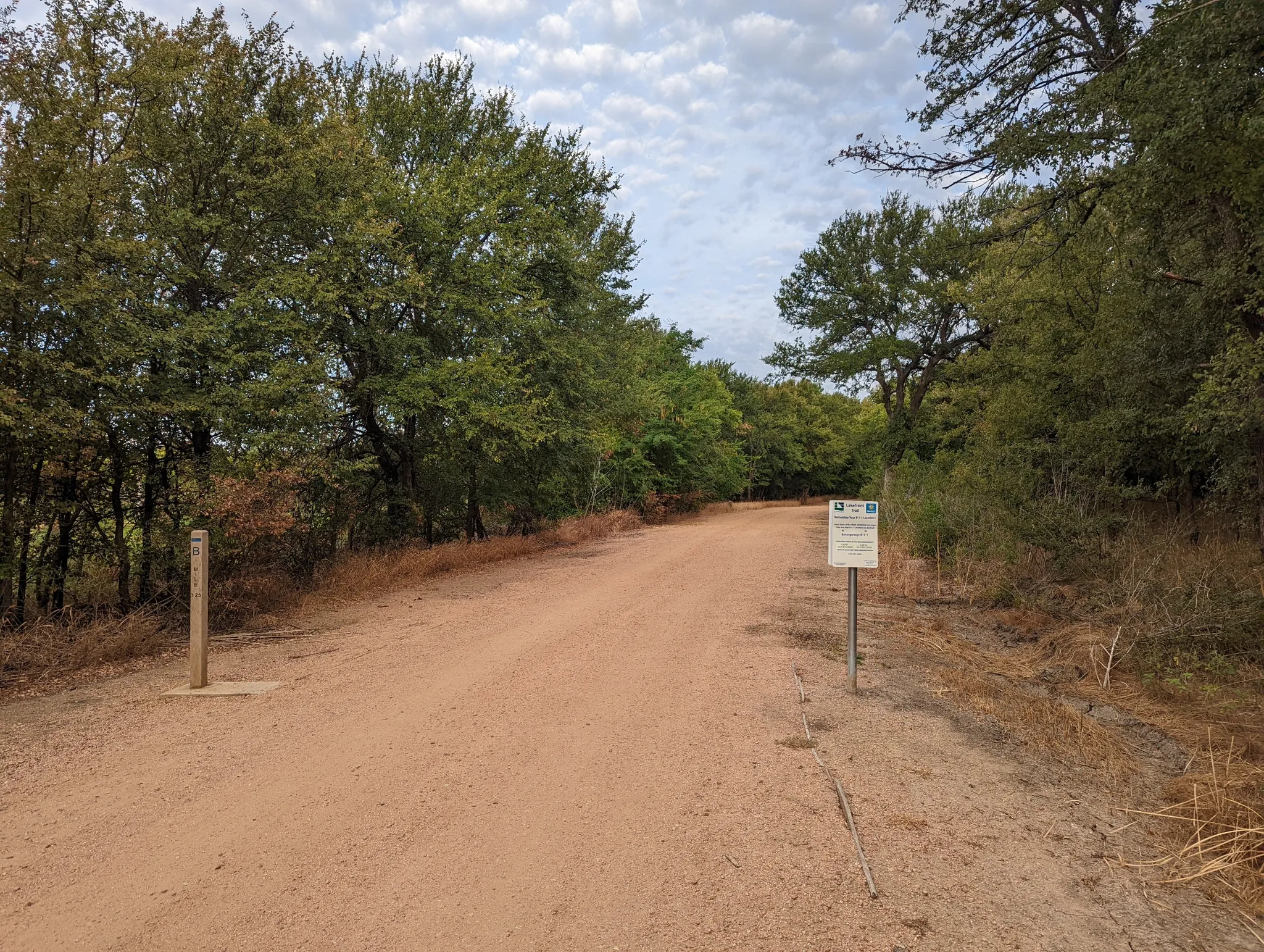 A wide dirt trail winding through dense green and brown foliage under a cloudy sky. - McCord Park — Gravel section – Little Elm Lakefront Trail loop from Bank of America