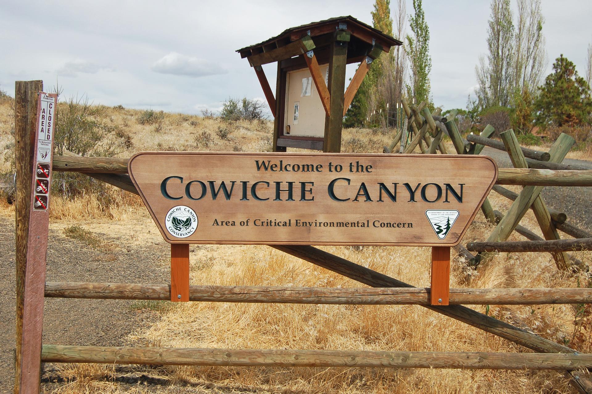 Wooden welcome sign on a fence in a dry, grassy landscape under an overcast sky.