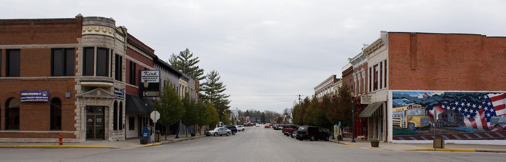 Empty town street lined with brick buildings and parked cars under overcast sky.