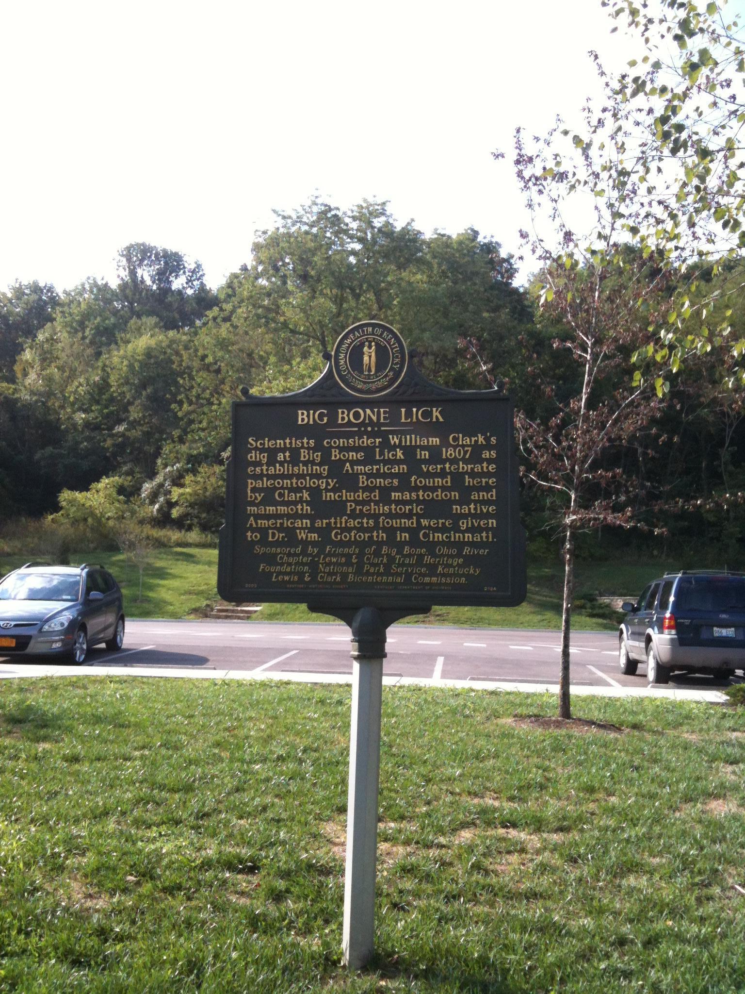 A historical marker stands in a grassy area with trees and cars.