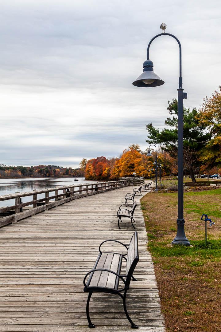 Wooden boardwalk with benches and a lamppost beside calm water, autumn trees.