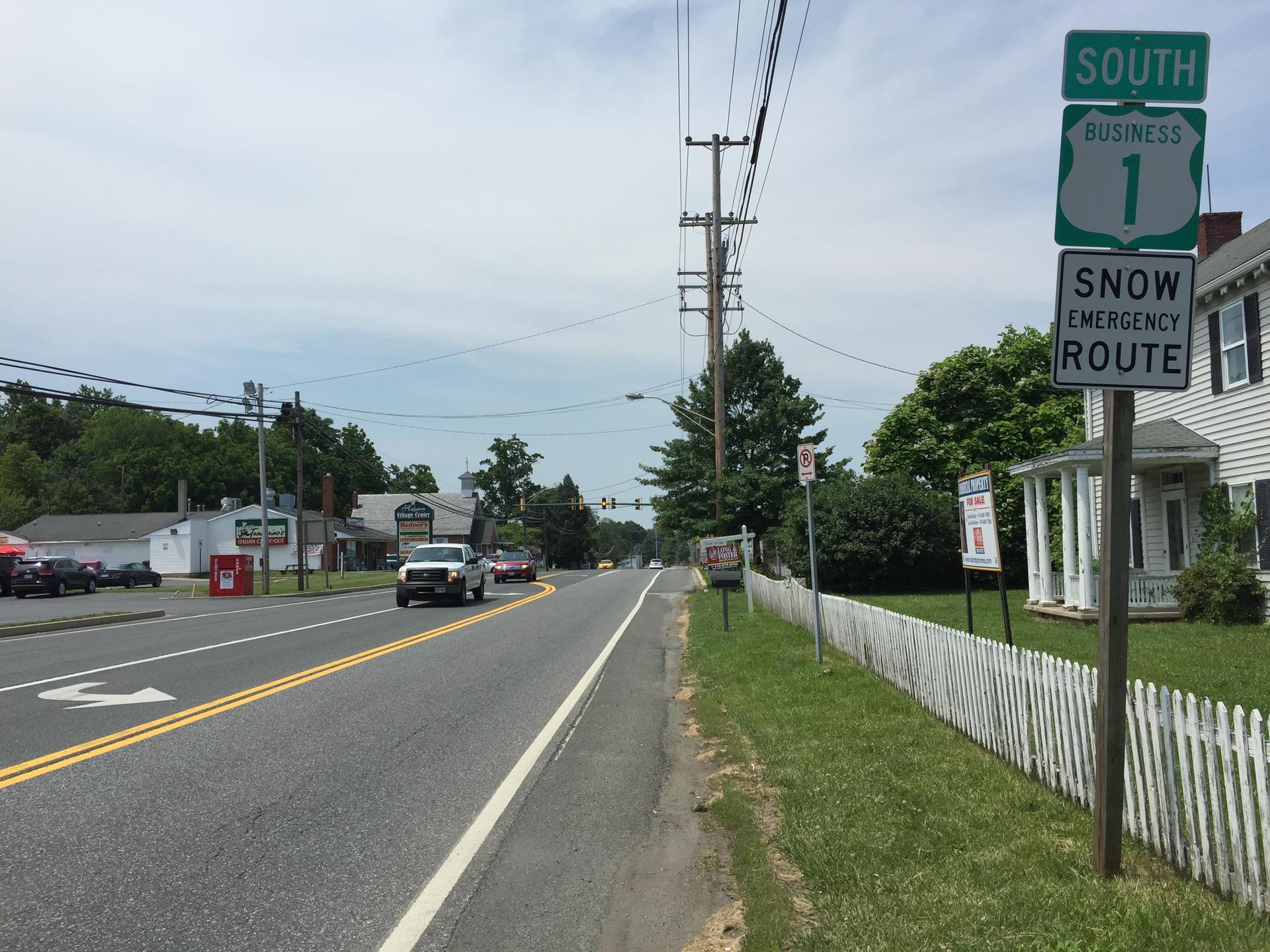 A suburban road with cars, power lines, and a white house under an overcast sky.