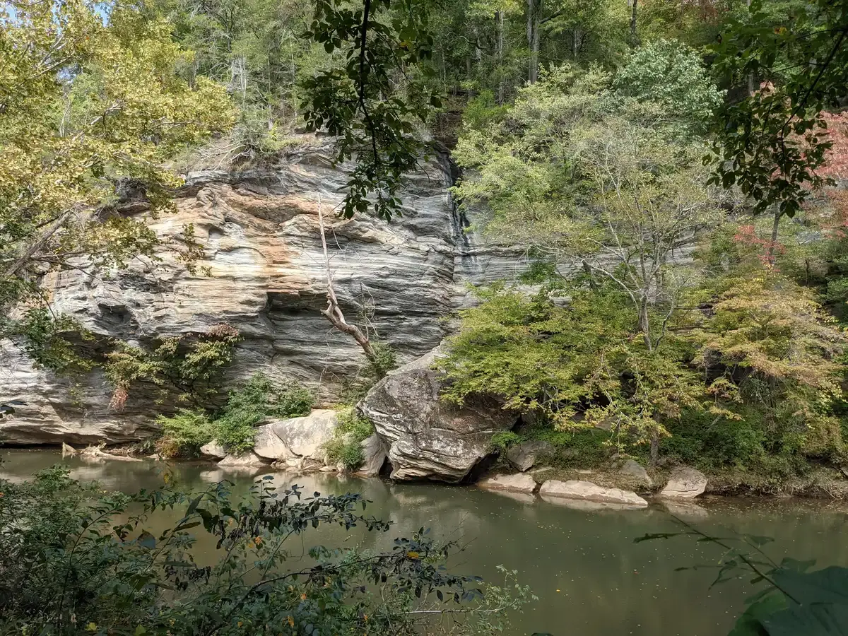 Calm river flowing past a large, layered rock face and green trees.