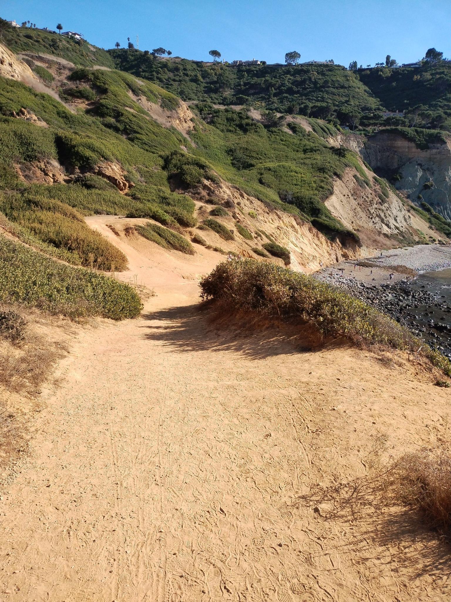 Sandy trail winding along a steep, green and brown hillside by the ocean.