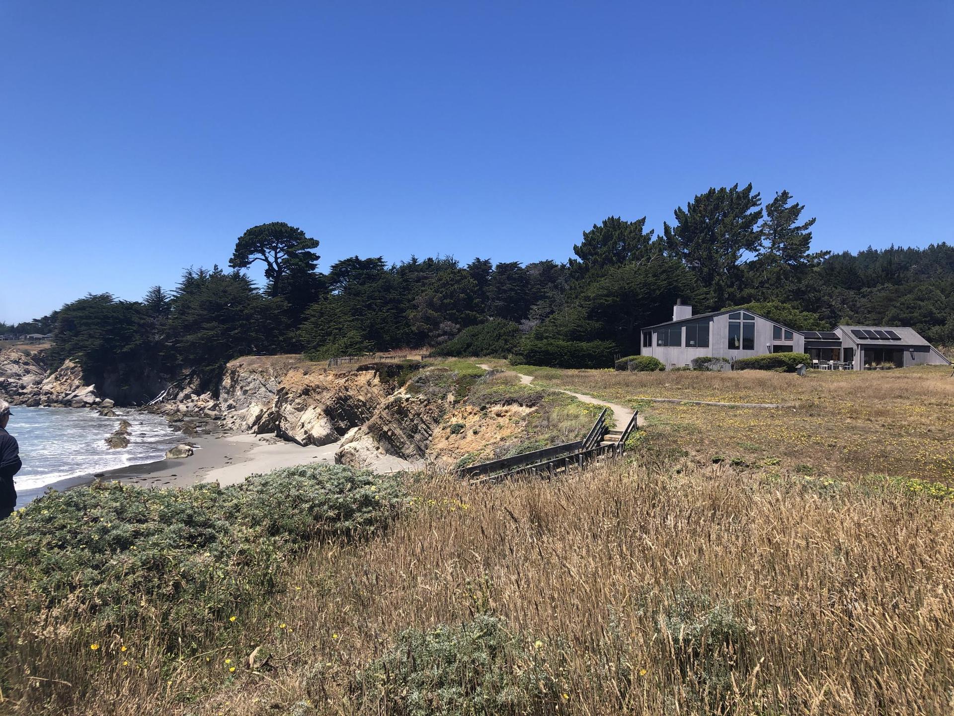 Coastal house on a grassy cliff overlooking a sunny beach and ocean.