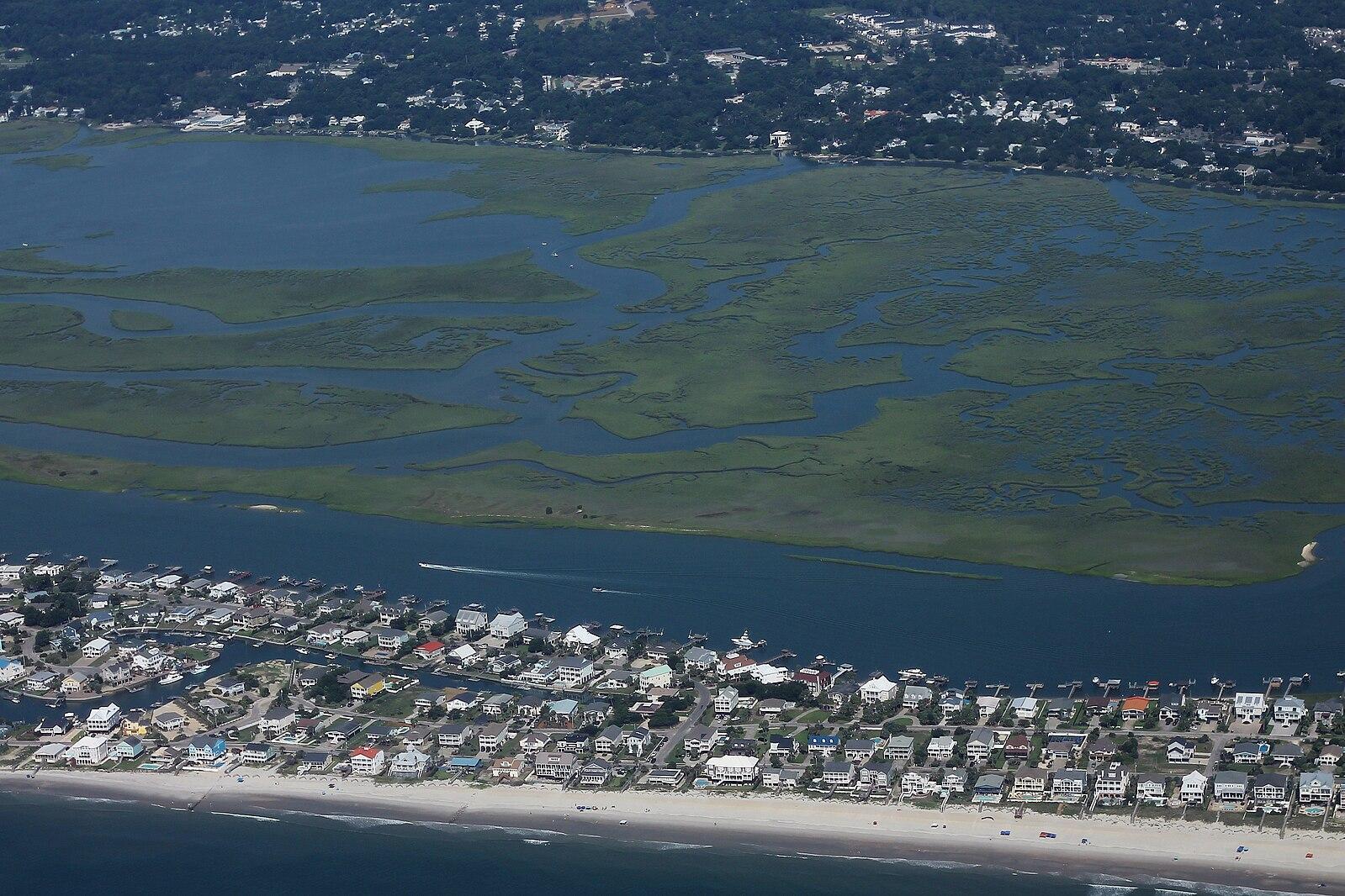 Aerial view of coastal homes, beach, marsh, and a boat on water.