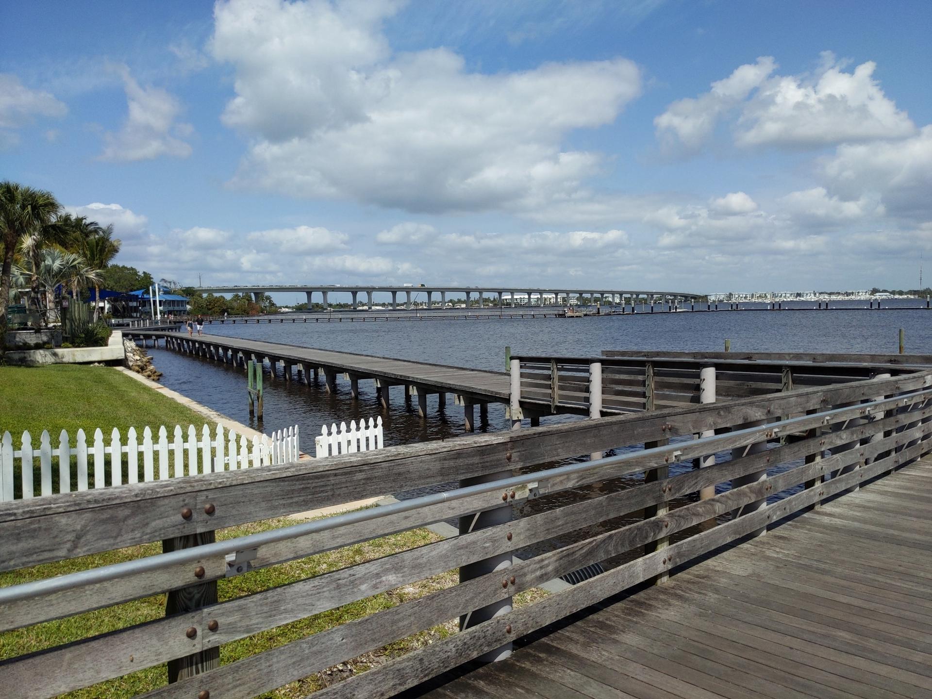A long wooden boardwalk and pier extend over calm water under a bright sky.
