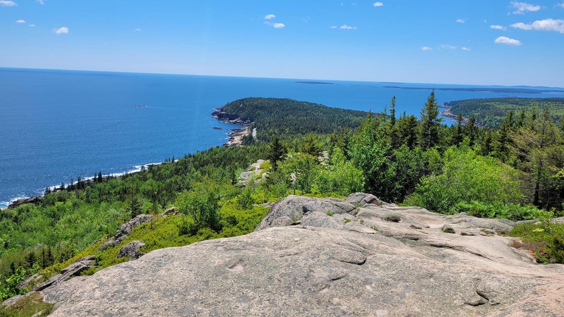 Sunny panoramic view from rocky peak, blue ocean, green forested coastline.