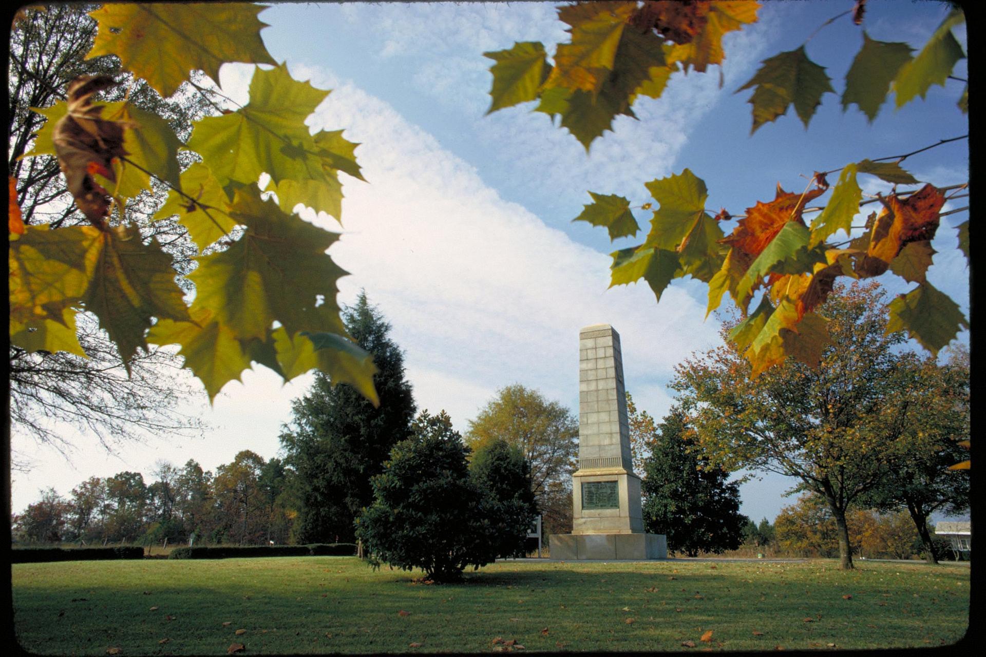 Stone obelisk monument in a park, framed by autumn leaves and trees.