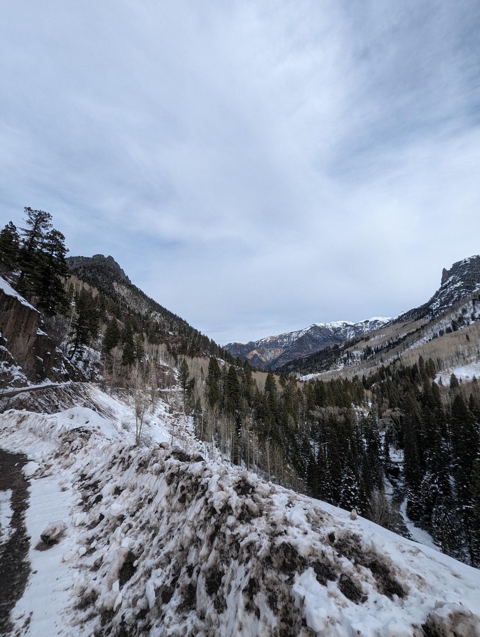 A snowy road winds through a winter mountain valley under an overcast sky.