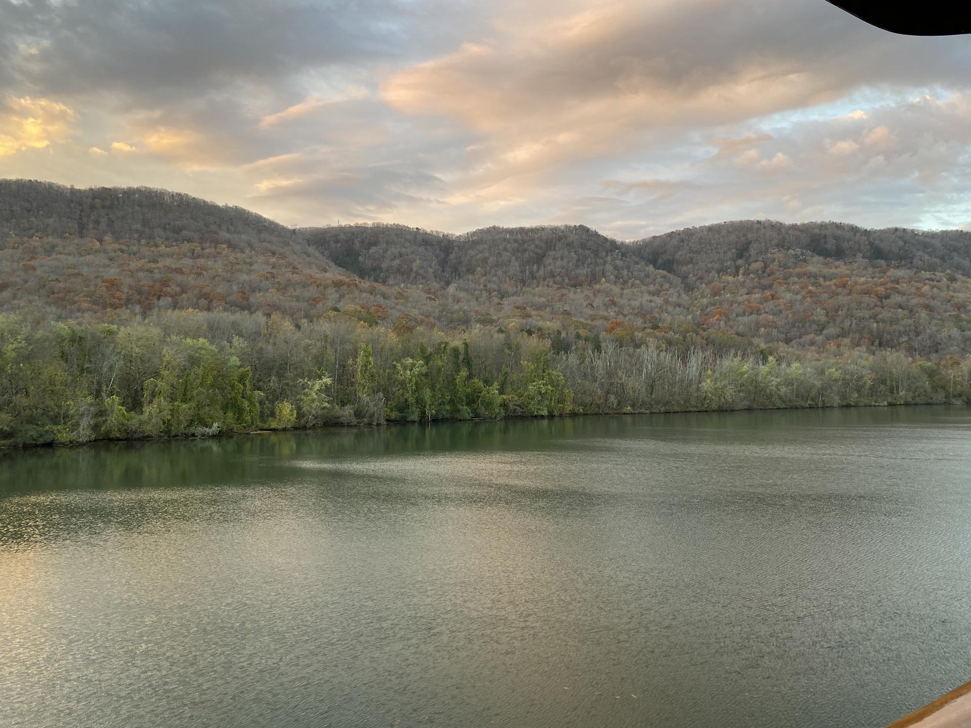 A wide river with forested hills under a cloudy, colorful sky at dusk.