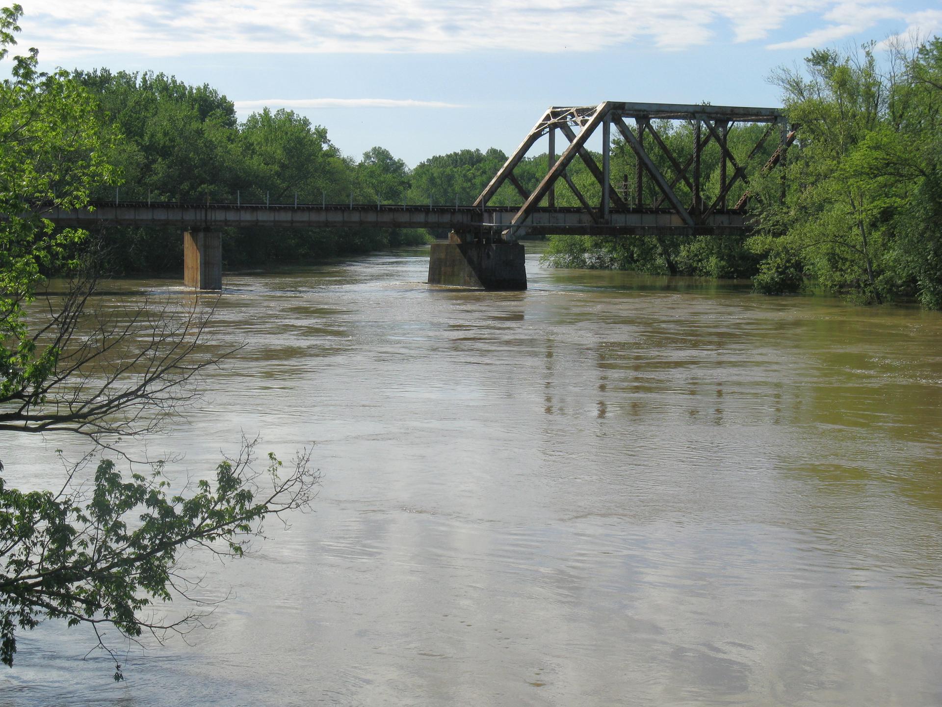 A metal truss bridge spans a wide, muddy river under a cloudy sky.