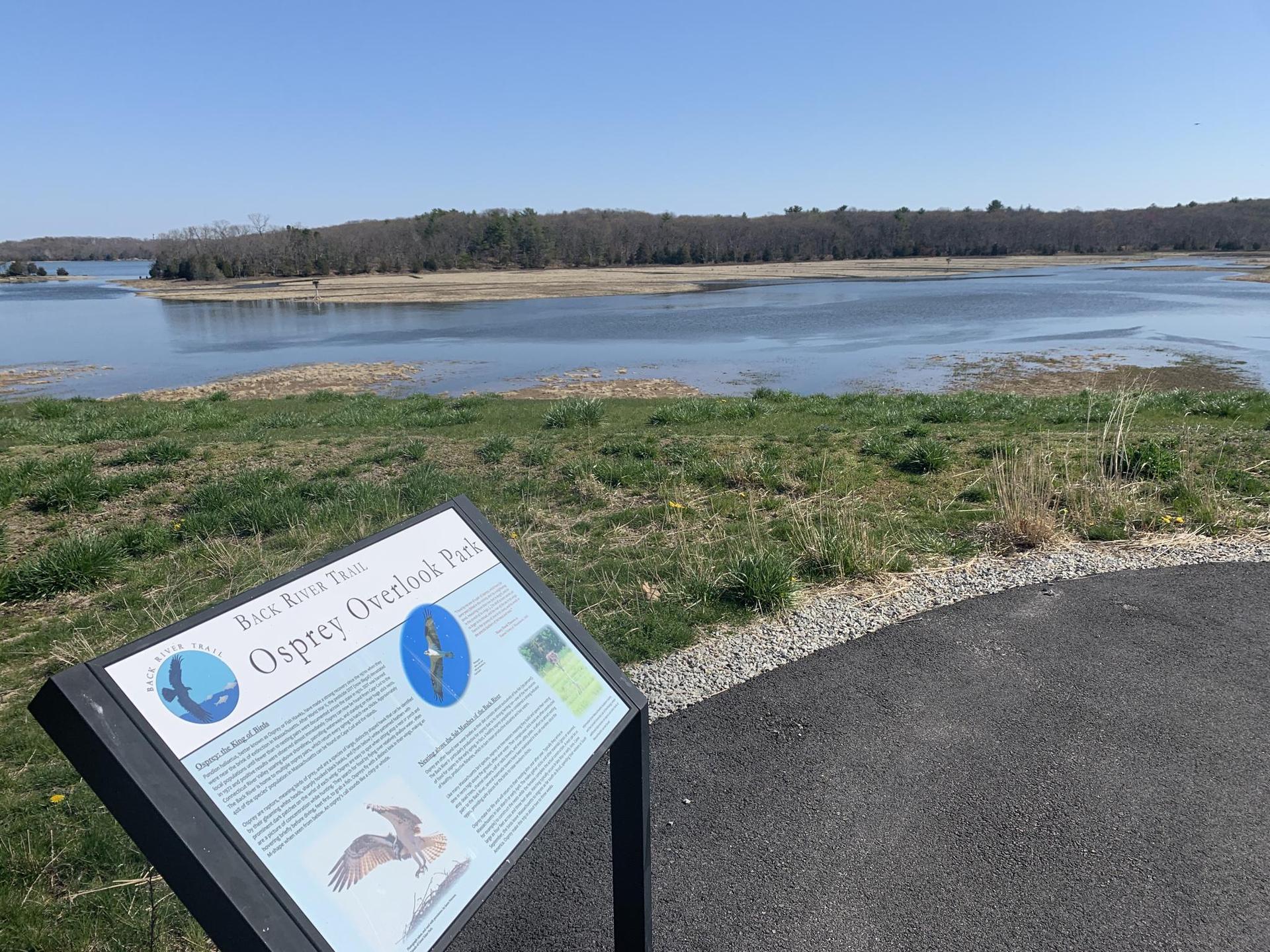 An informational sign in the foreground, with a wide river and trees beyond.
