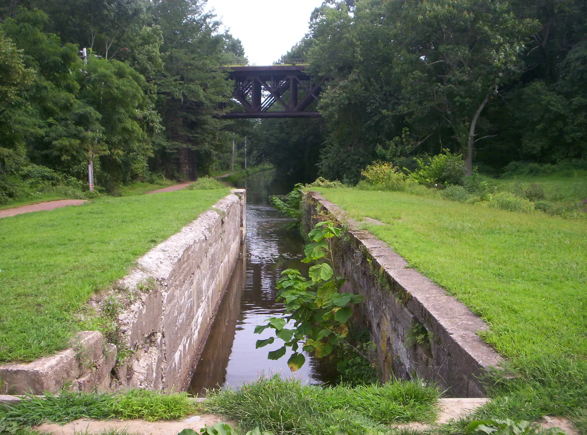 Historic canal lock with stone walls and a metal bridge overhead, surrounded by green.