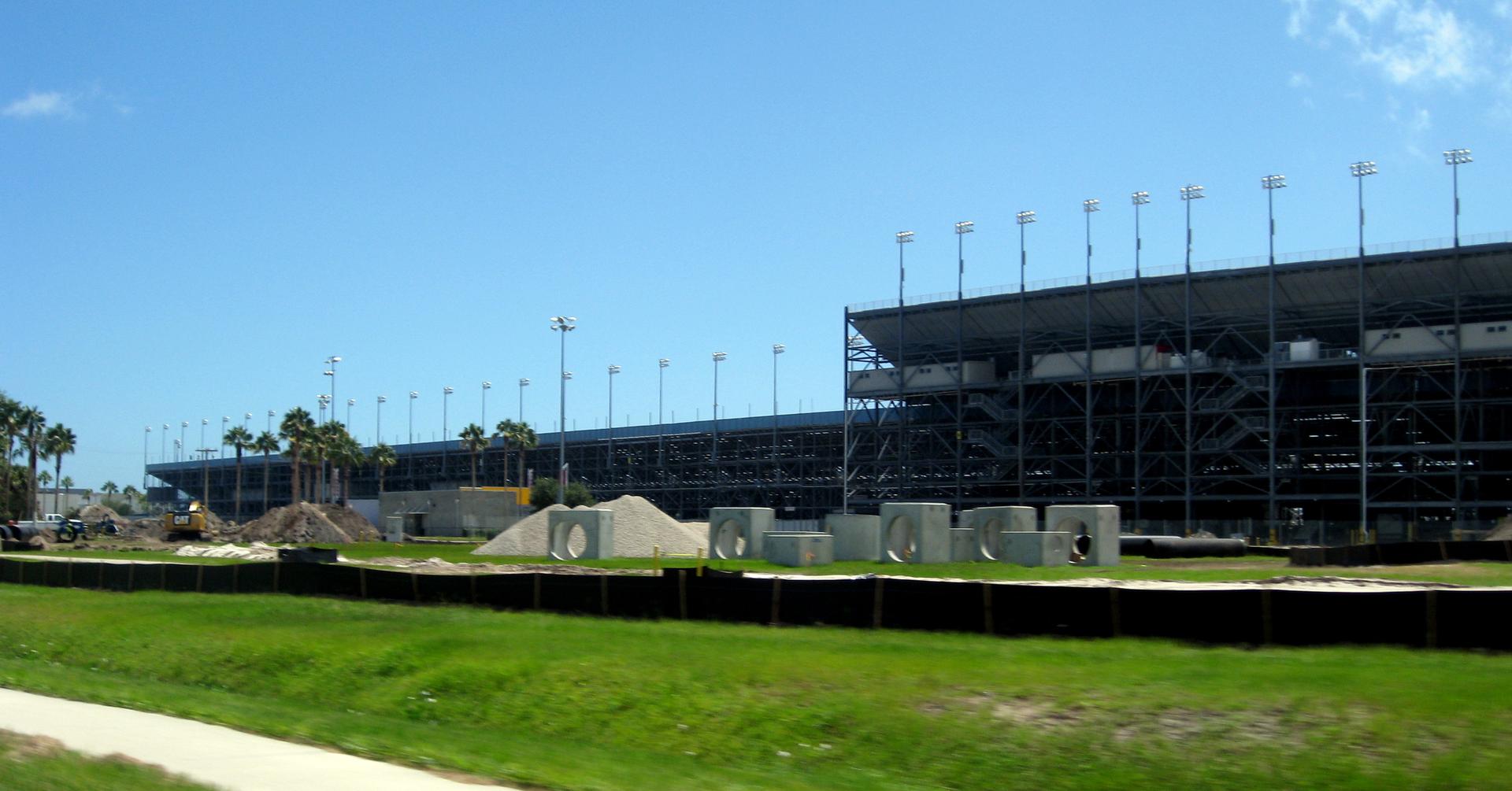 Large stadium grandstands under construction with palm trees under a bright blue sky.