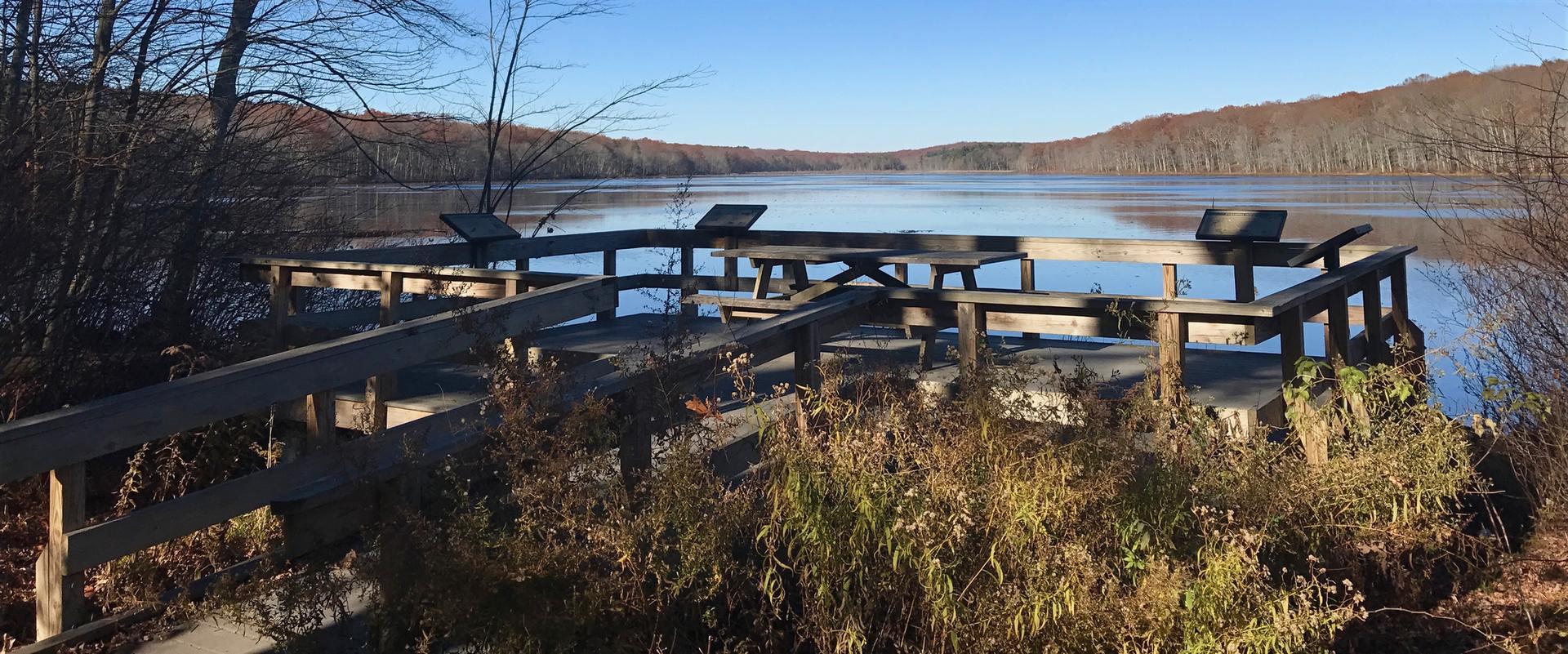 A wooden viewing platform overlooks a calm lake and distant forested hills.