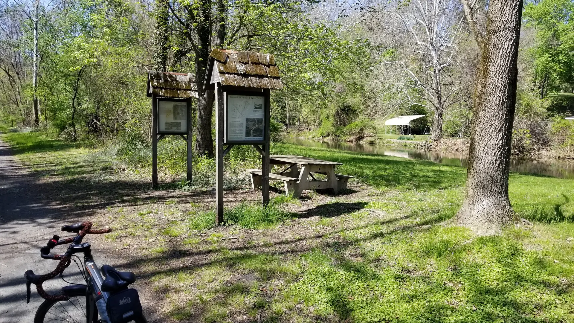 Bicycle parked near picnic table and signs by calm water, sunny day.