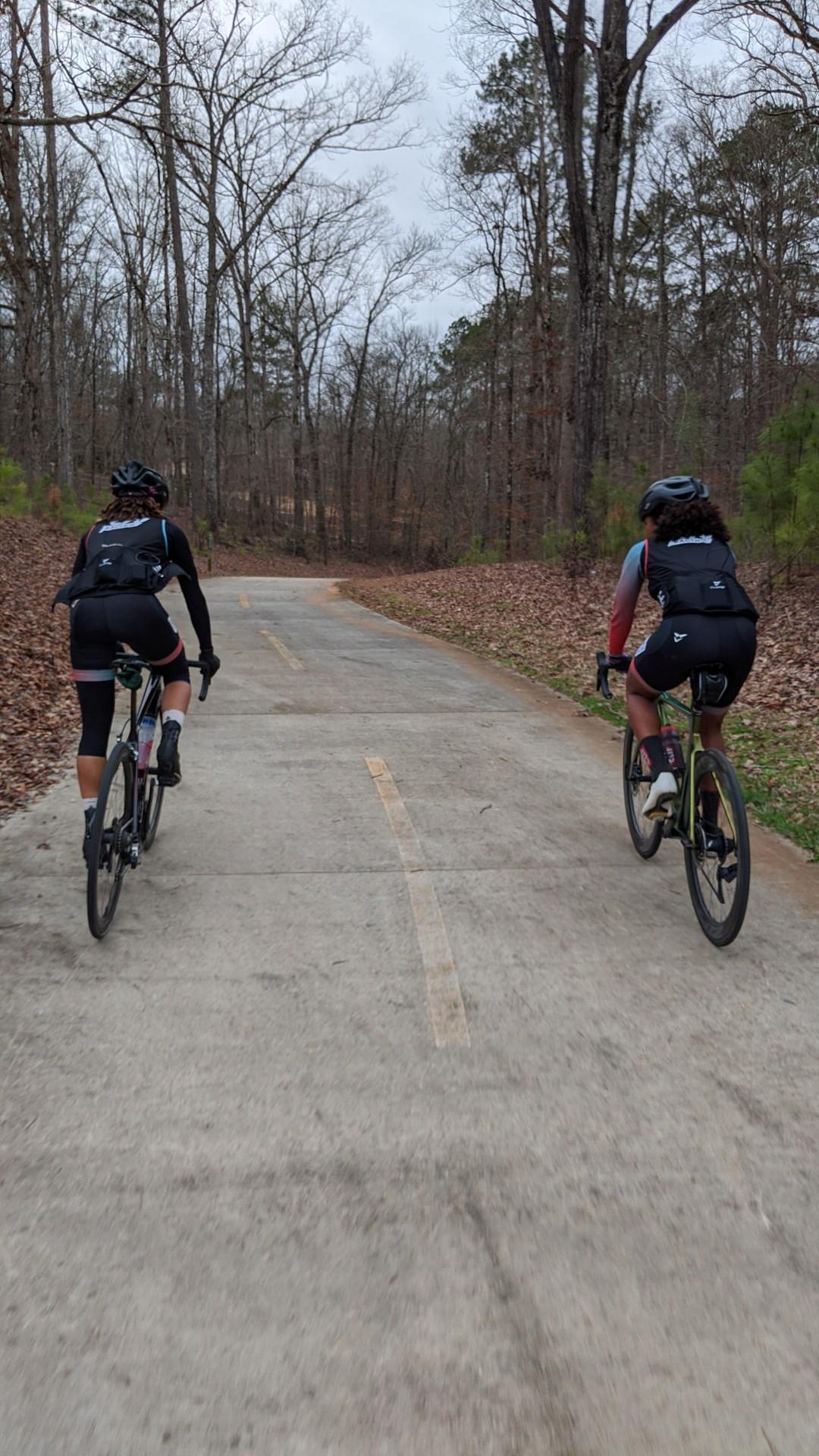 Two cyclists ride away on a paved path through a forest with bare trees.