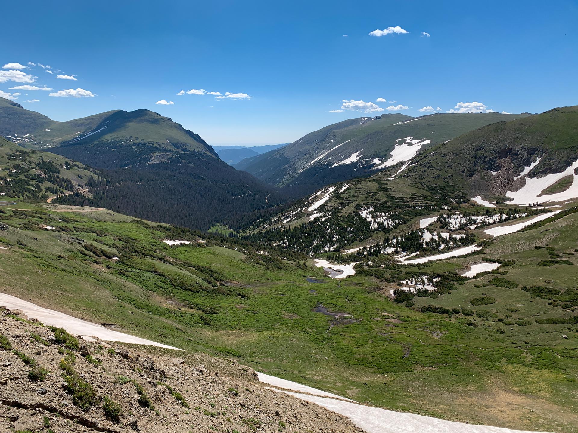 A vast mountain valley with green slopes, patches of snow, under a blue sky.