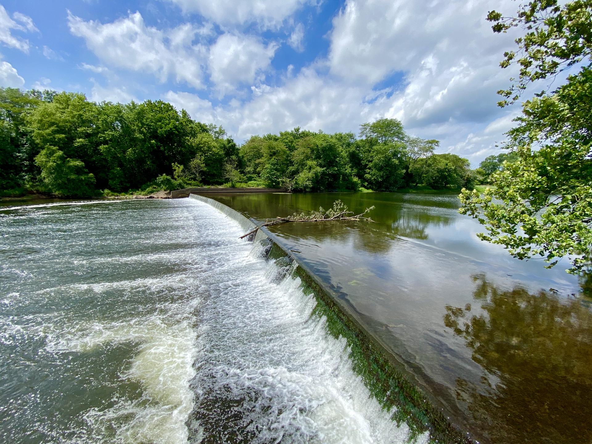 Sunny river scene with water flowing over a dam, lush green trees, and blue sky.