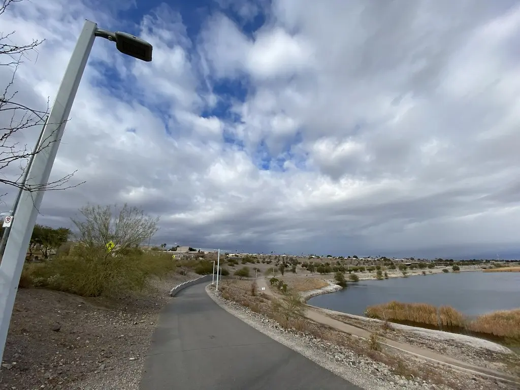 Paved path curving beside a lake under a cloudy sky with a street light.