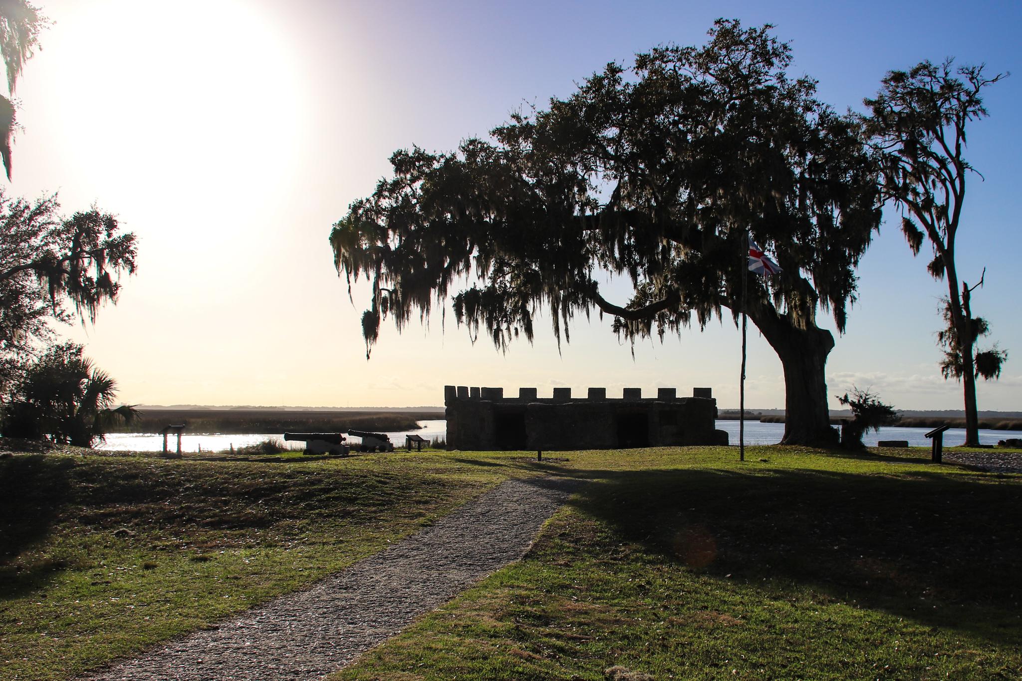 Sunny view of a stone fort and moss-draped trees by calm water.