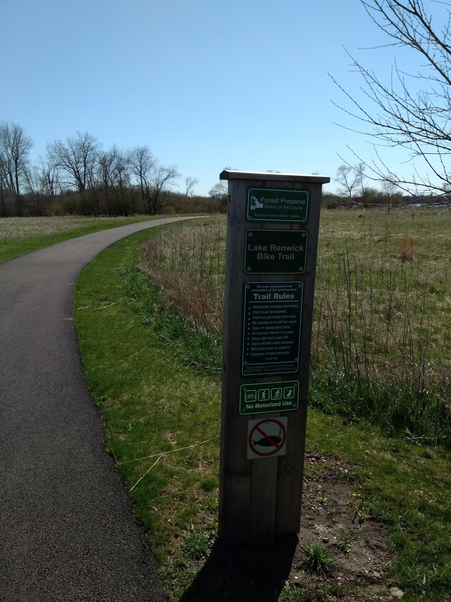 Sunny view of a trail sign beside a paved path and grassy field.