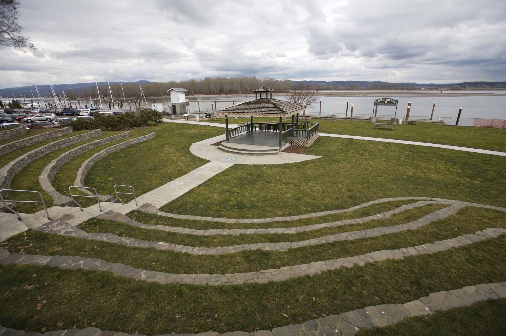 A waterfront park with a gazebo, amphitheater seating, and a marina under cloudy skies.