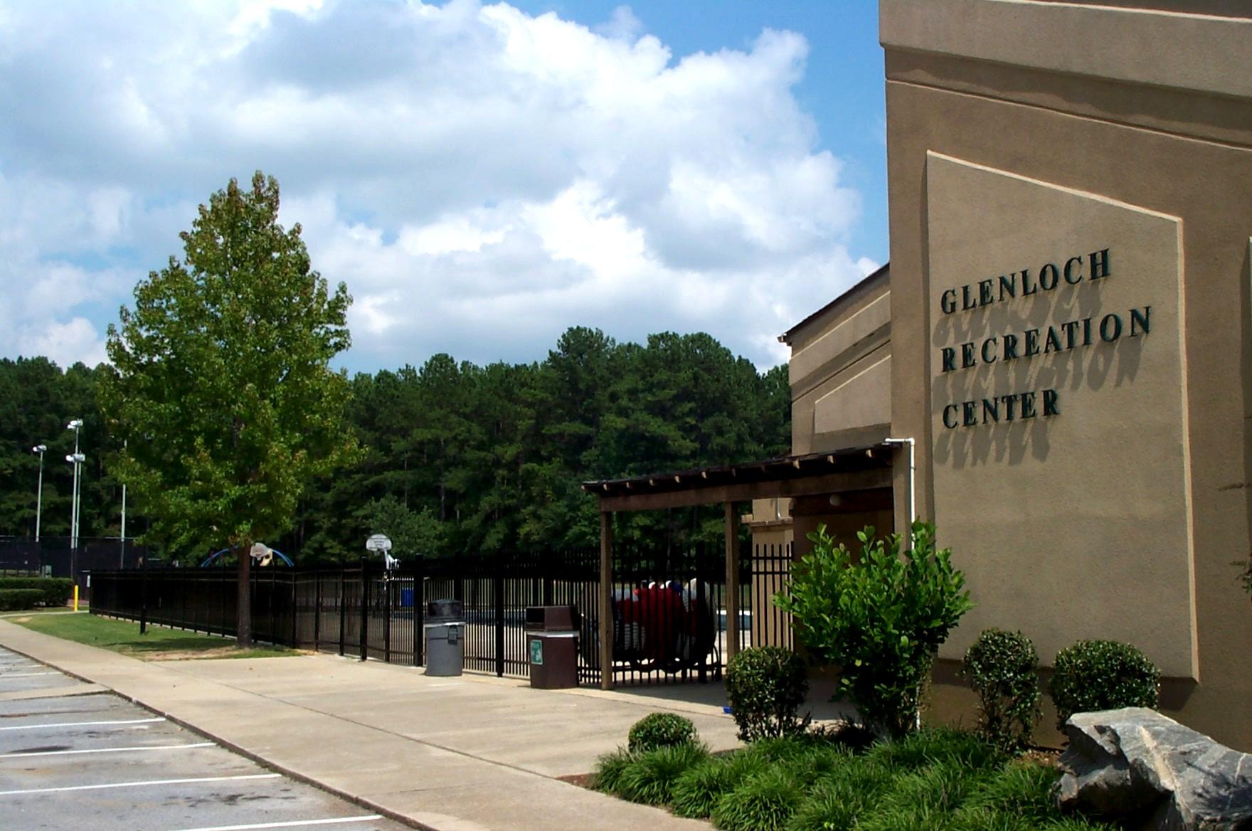Building exterior with a sign, paved area, and green trees under a cloudy sky.
