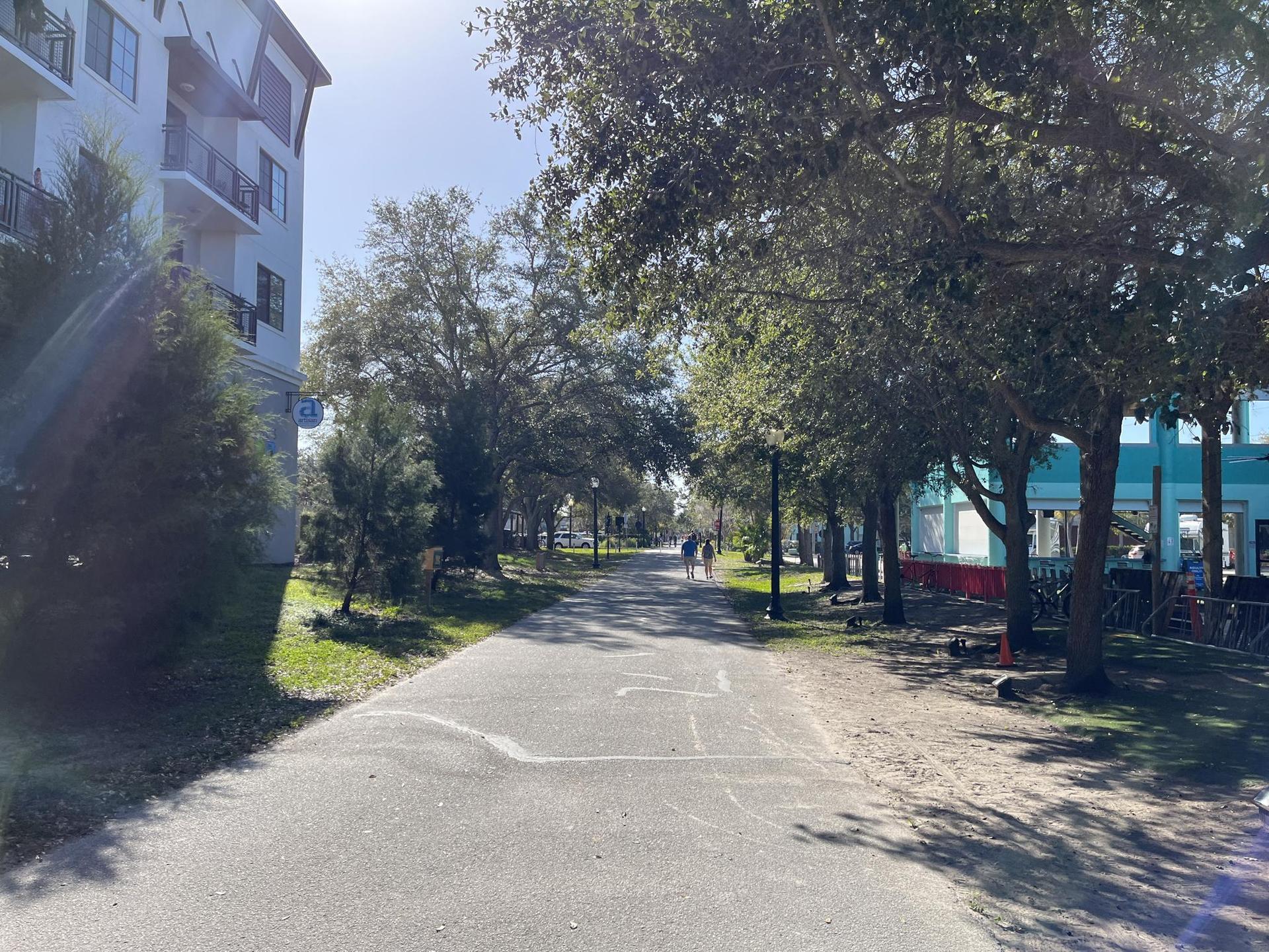 Sunny paved path lined with trees and buildings, two people walking.