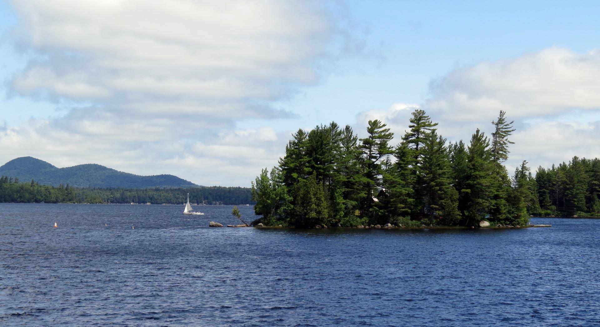 Daylight view of a small island in a lake with a sailboat and distant mountains.