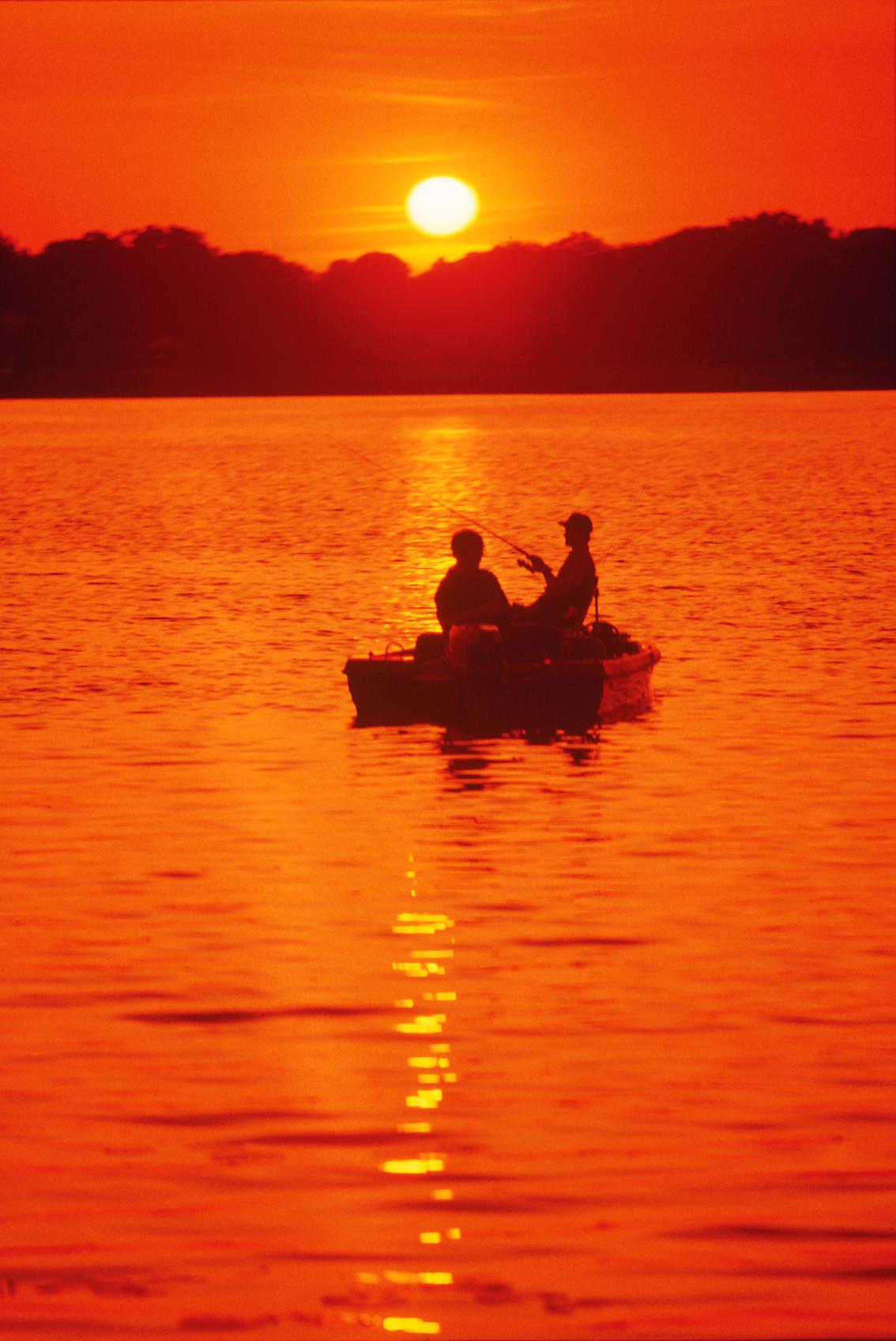 Two people in a boat silhouetted against a vibrant orange sunset over water.