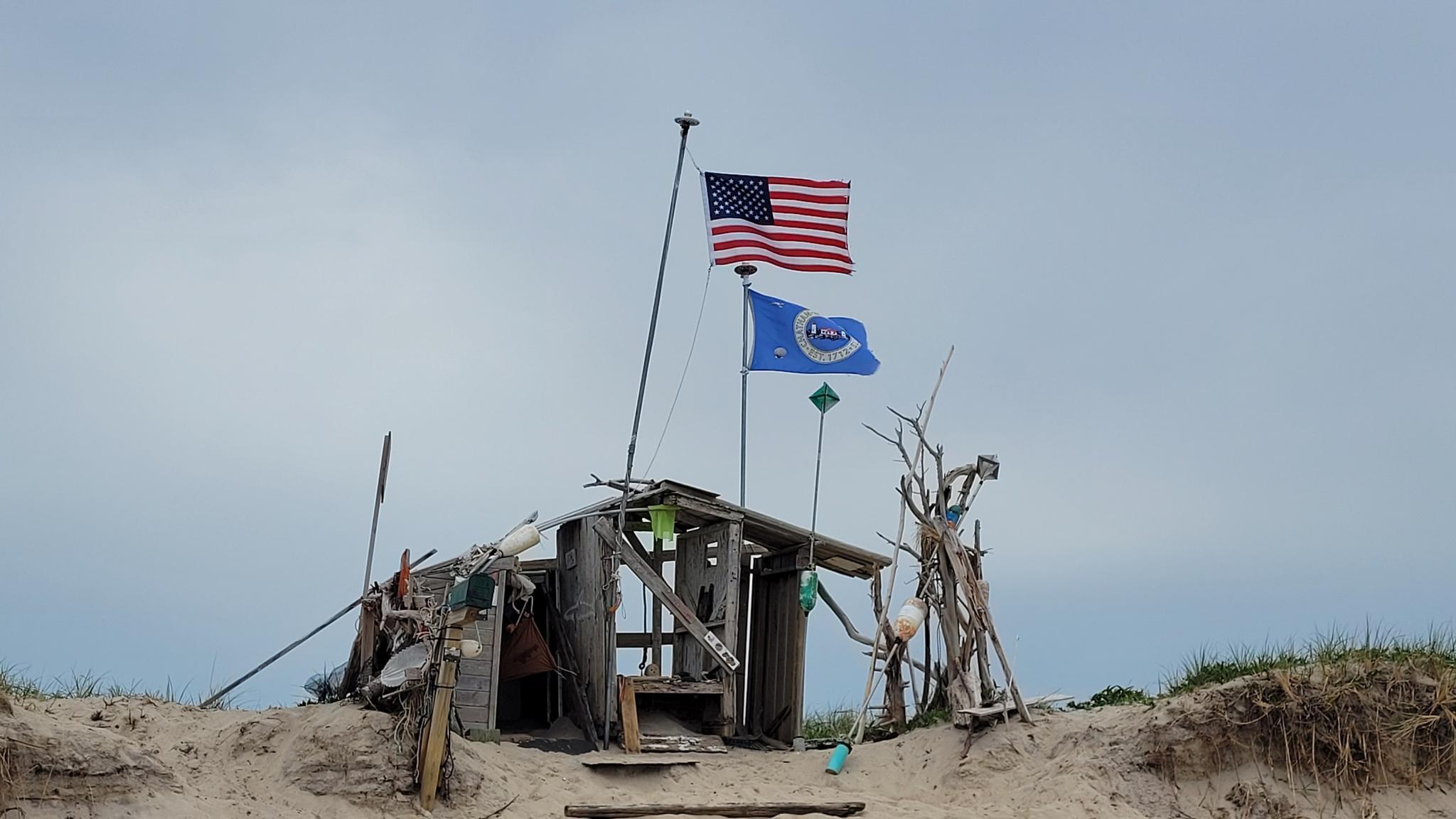 Rustic shack on a sand dune with two flags flying under a cloudy sky.