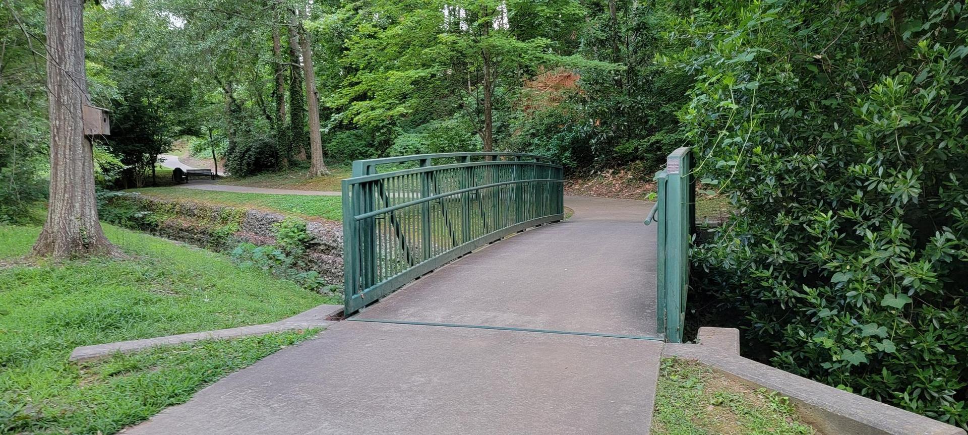 Green metal bridge over a narrow stream in a lush, green park.