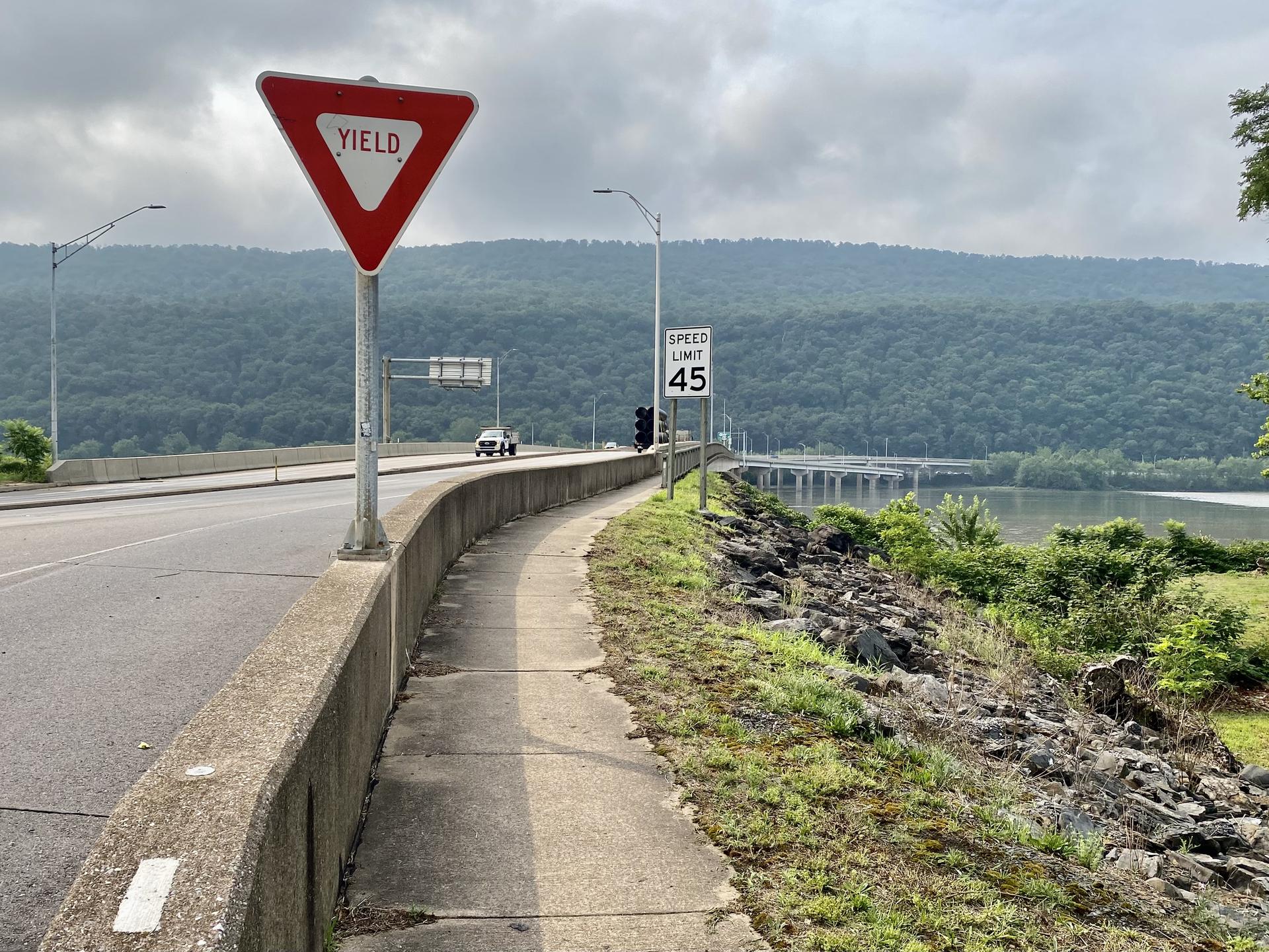 Roadside view with yield sign, river, and forested mountains under an overcast sky.