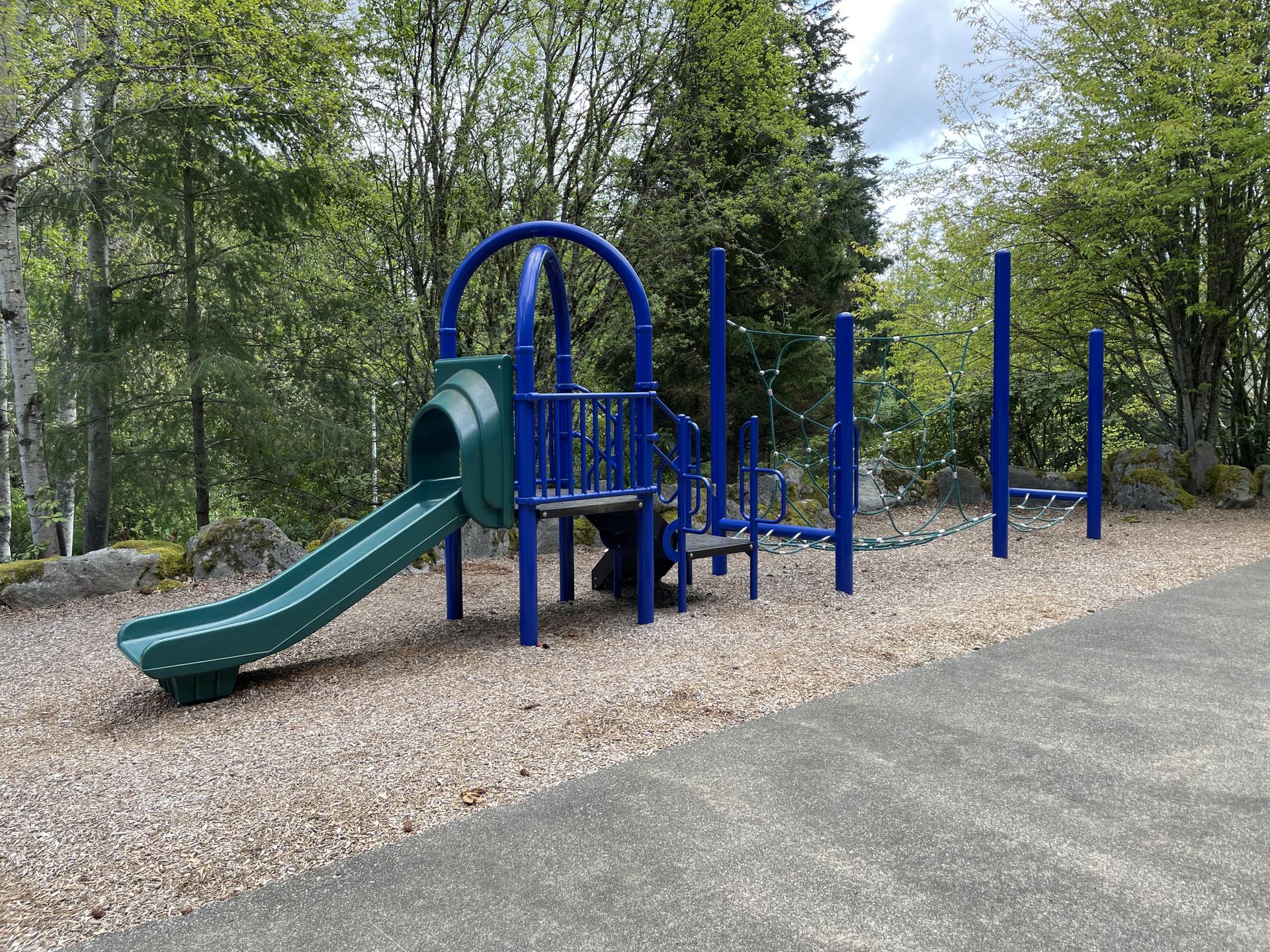 A blue and green playground with a slide and climbing nets, surrounded by green trees.