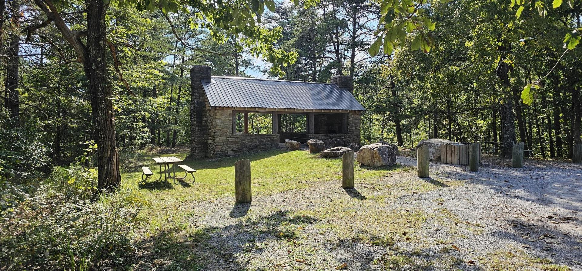 A stone pavilion with a metal roof sits in a sunny forest clearing.