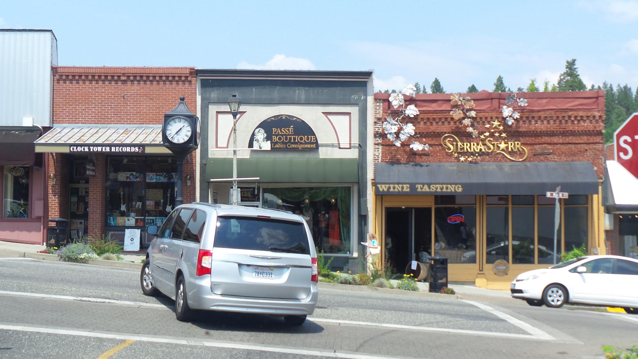 A street scene with multiple storefronts, parked cars, and a clock in bright daylight.