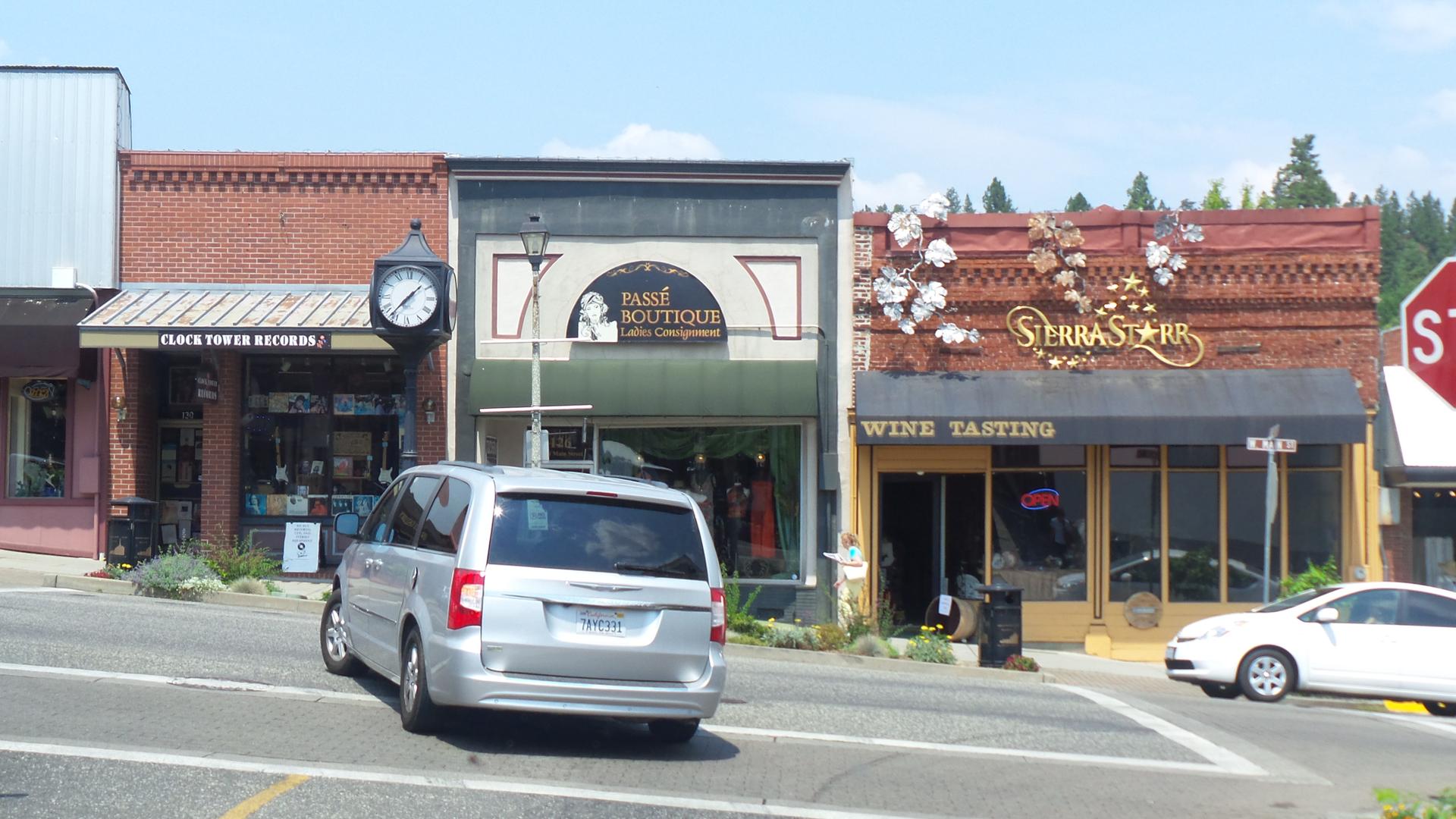 A street scene with multiple storefronts, parked cars, and a clock in bright daylight.