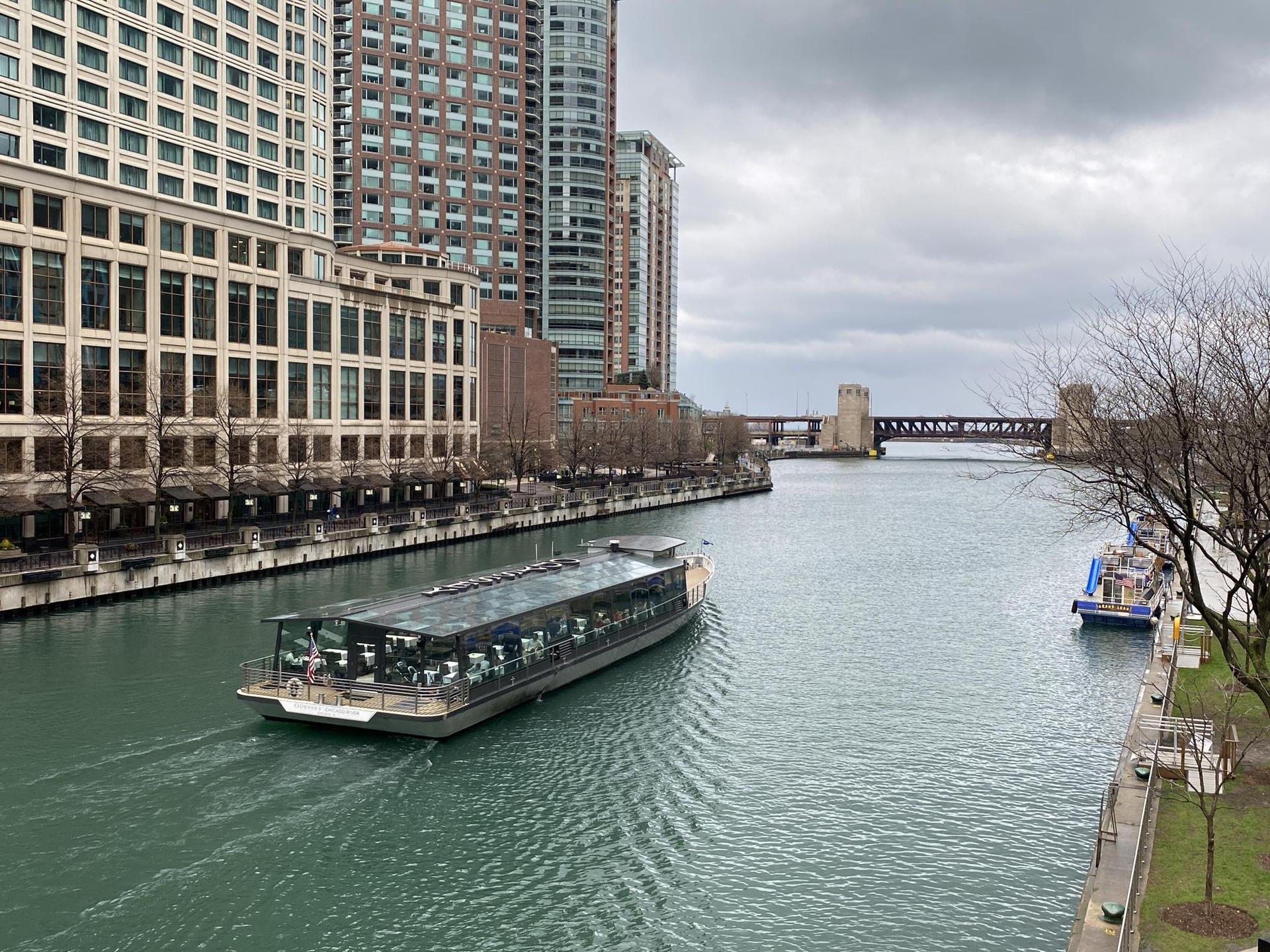 A tour boat on a river, surrounded by tall city buildings on a cloudy day.