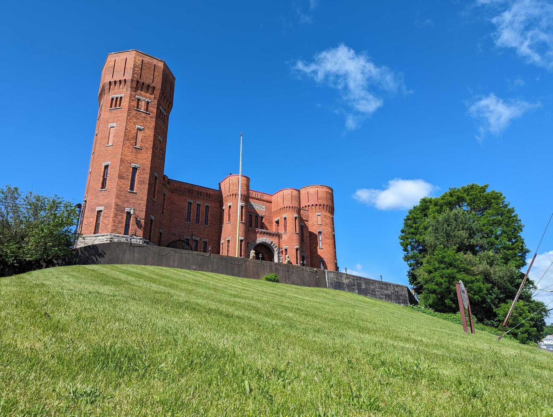 Large brick building with towers on a grassy hill under a bright blue sky.