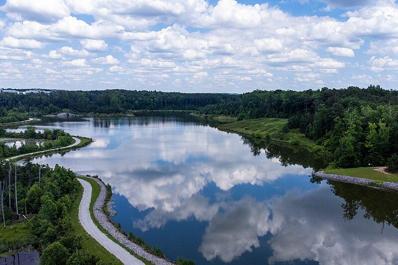 Aerial view of a lake reflecting clouds, bordered by green forests and a winding path.