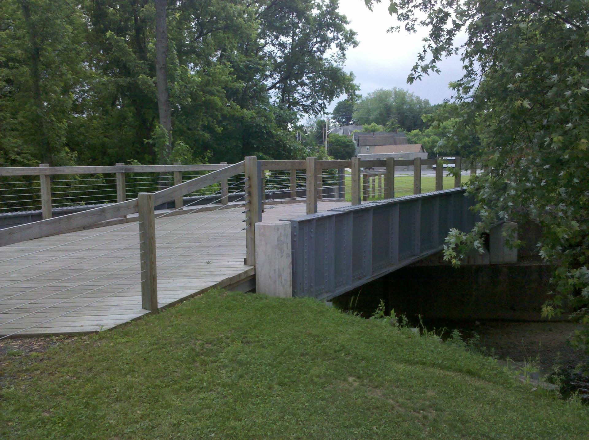 A modern wooden and metal pedestrian bridge over water, surrounded by trees.