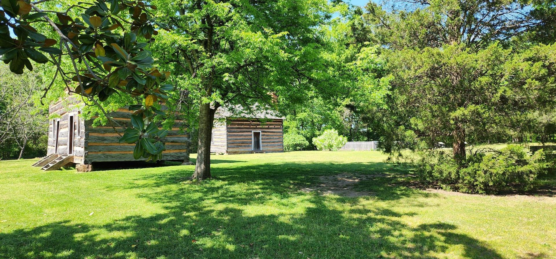Two rustic wooden cabins in a sunny grassy field with trees.