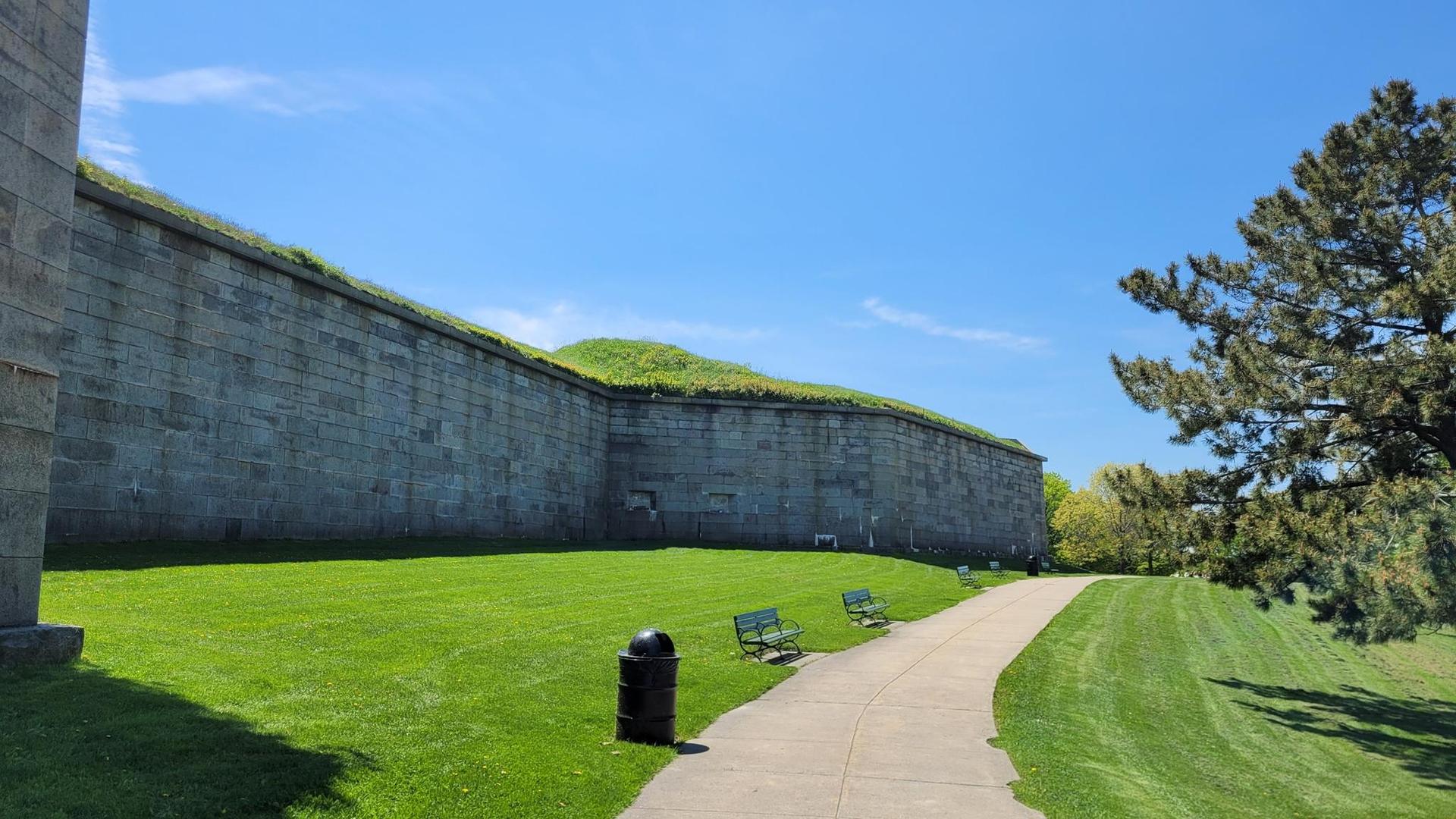Massive stone wall with grassy top, green lawn, paved path, and trees under clear sky.