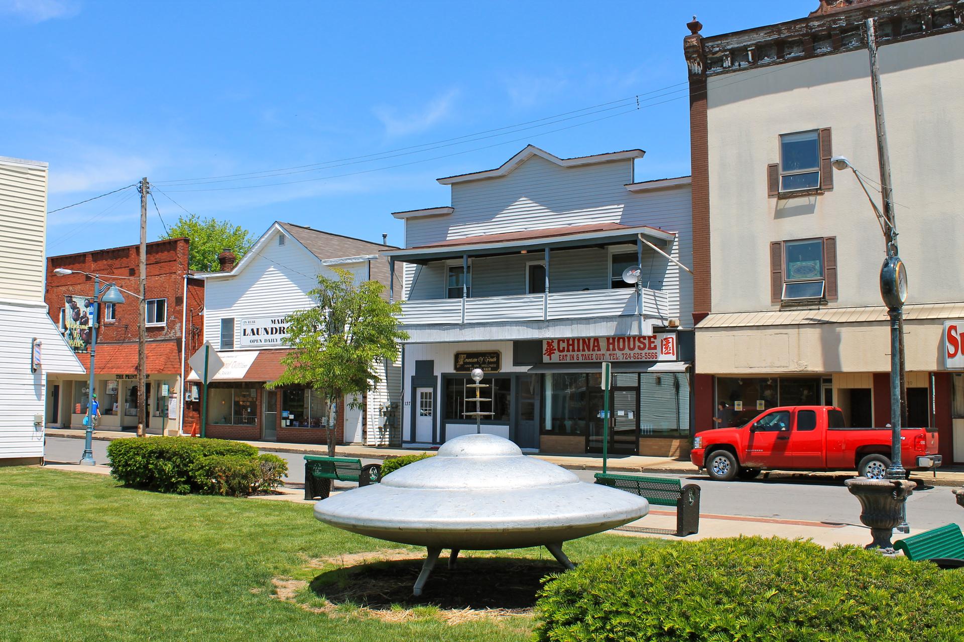 A town square with a silver UFO sculpture, buildings, and a red truck.